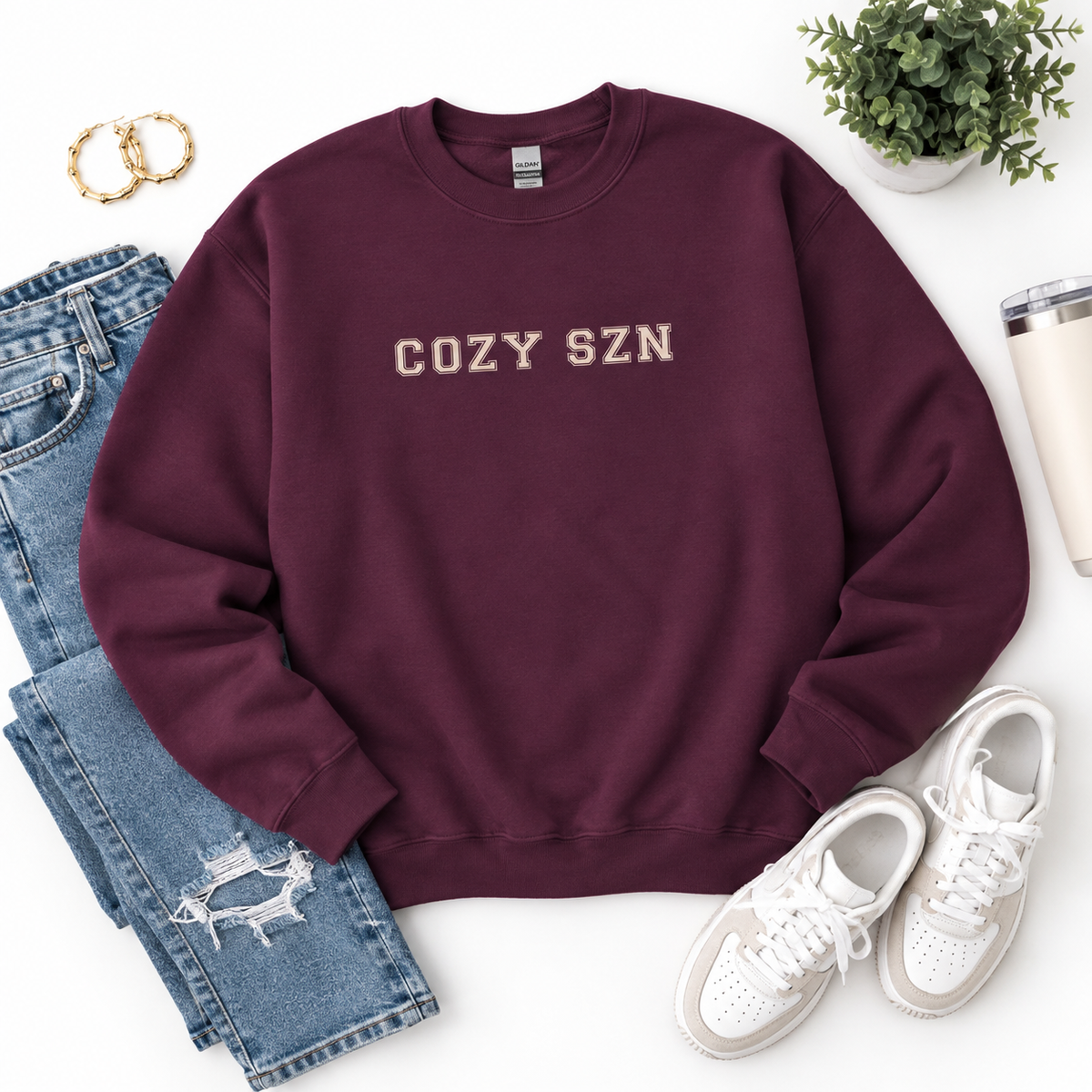A maroon crewneck sweatshirt with 'COZY SZN' printed in beige across the chest, styled in a flat lay with blue jeans, white sneakers, gold hoop earrings, a small potted plant, and a travel tumbler on a white background.