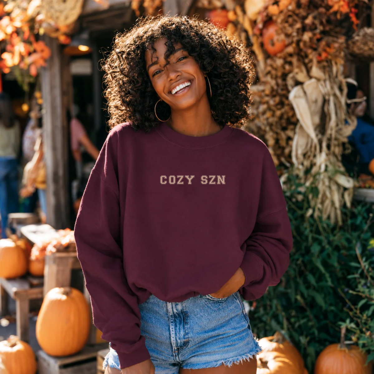 A woman with curly hair stands outdoors in a fall market setting wearing a maroon crewneck sweatshirt with 'COZY SZN' printed in beige across the chest, paired with denim shorts.